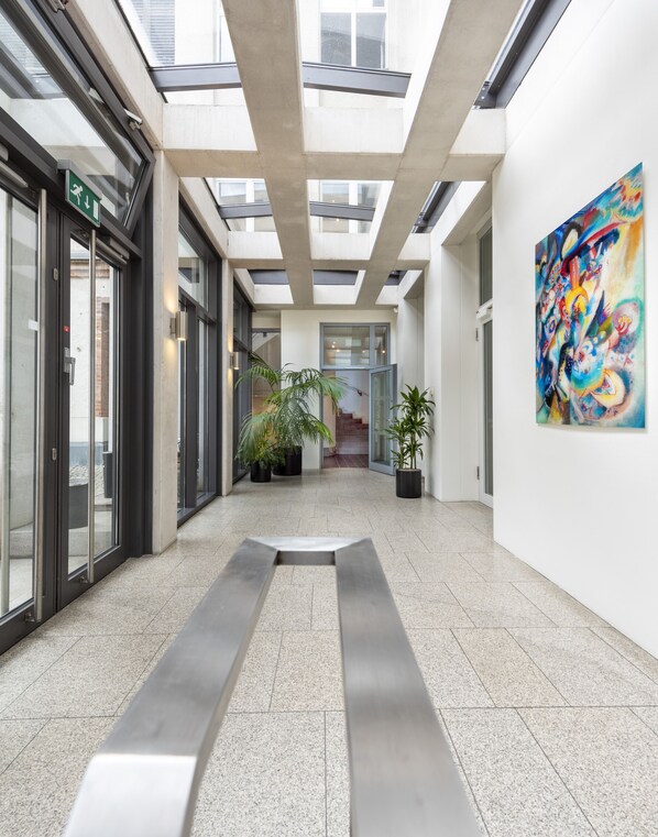 A modern interior with a central walkway, glass doors, potted plants, and a colorful abstract painting on the wall. - Hotel Aquino Berlin (Berlin)