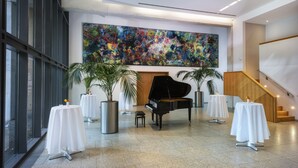 Un vestíbulo espacioso con un piano de cola, mesas blancas, plantas en macetas y un cuadro abstracto grande en la pared.