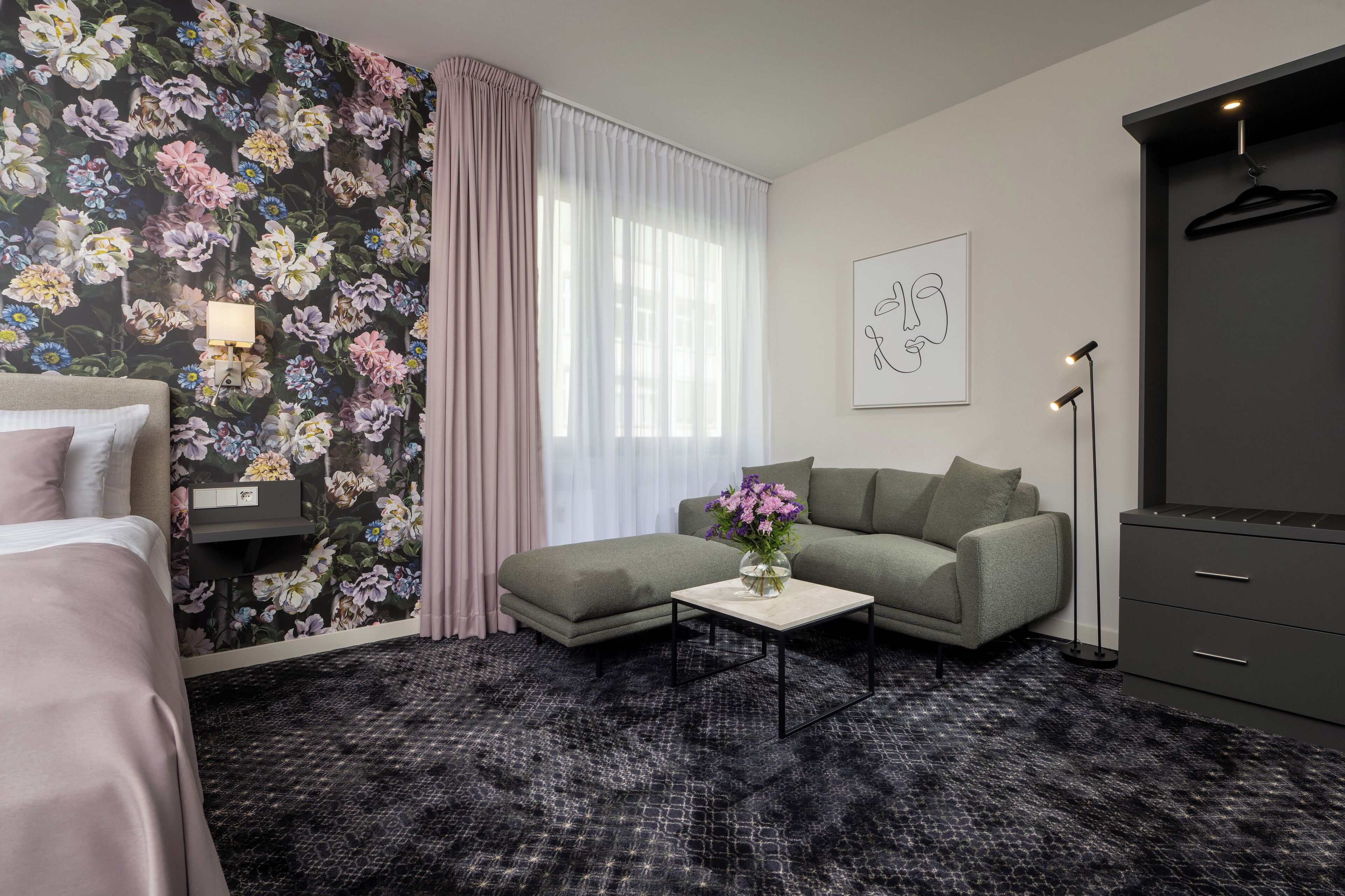 A hotel room with a bed, a sofa, a small table with flowers, a wardrobe, and a painting on the wall.
