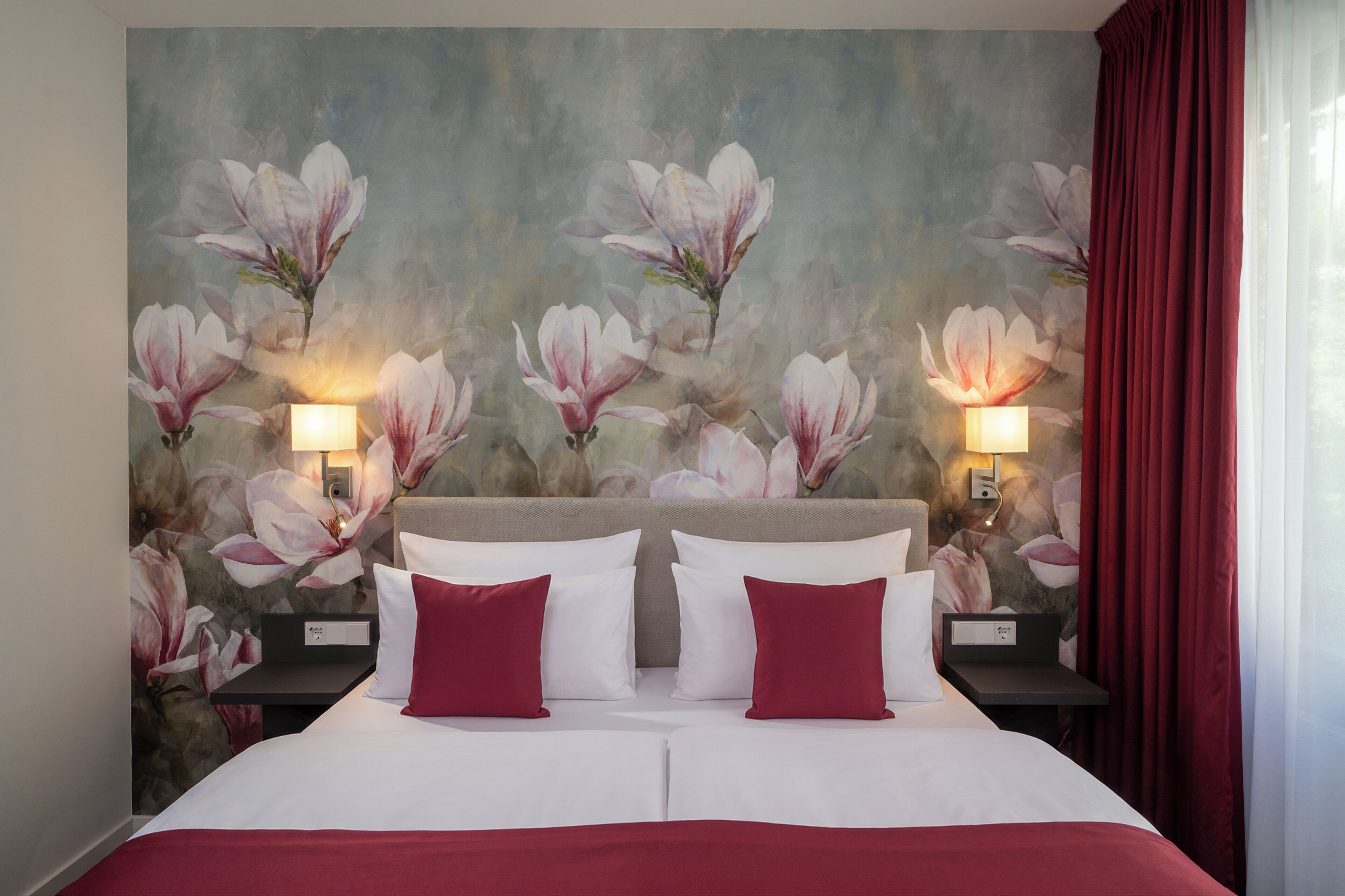 Una cama doble con ropa de cama blanca y roja, dos lámparas de noche y un mural floral en la pared.