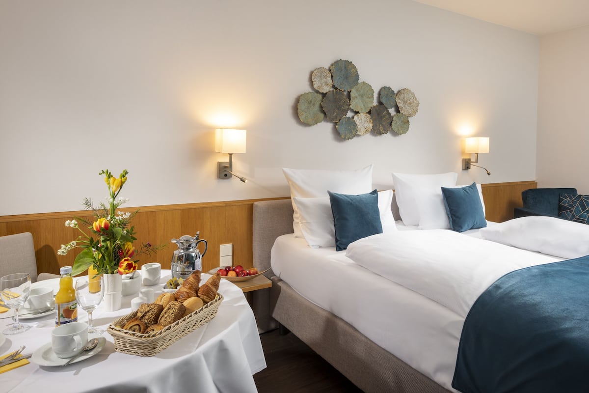Una habitación de hotel con una cama bien hecha, una mesa de desayuno con croissants y fruta, y un adorno en la pared.