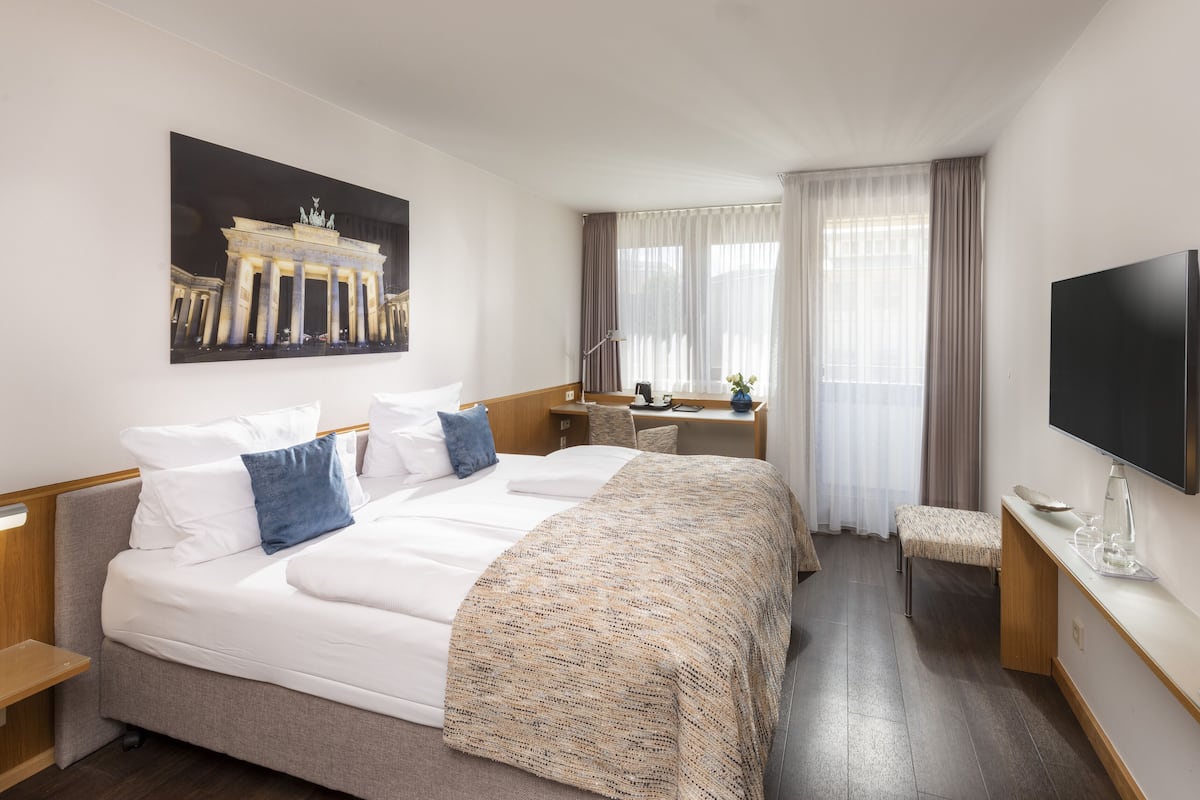 Habitación de hotel con una cama grande, un escritorio con silla, un televisor de pantalla plana y un cuadro de un edificio.
