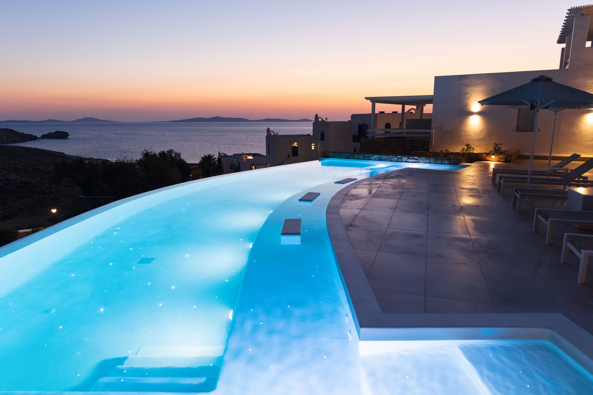 Una piscina con vista al mar al anochecer, rodeada de sillas de descanso y un edificio con zona cubierta.