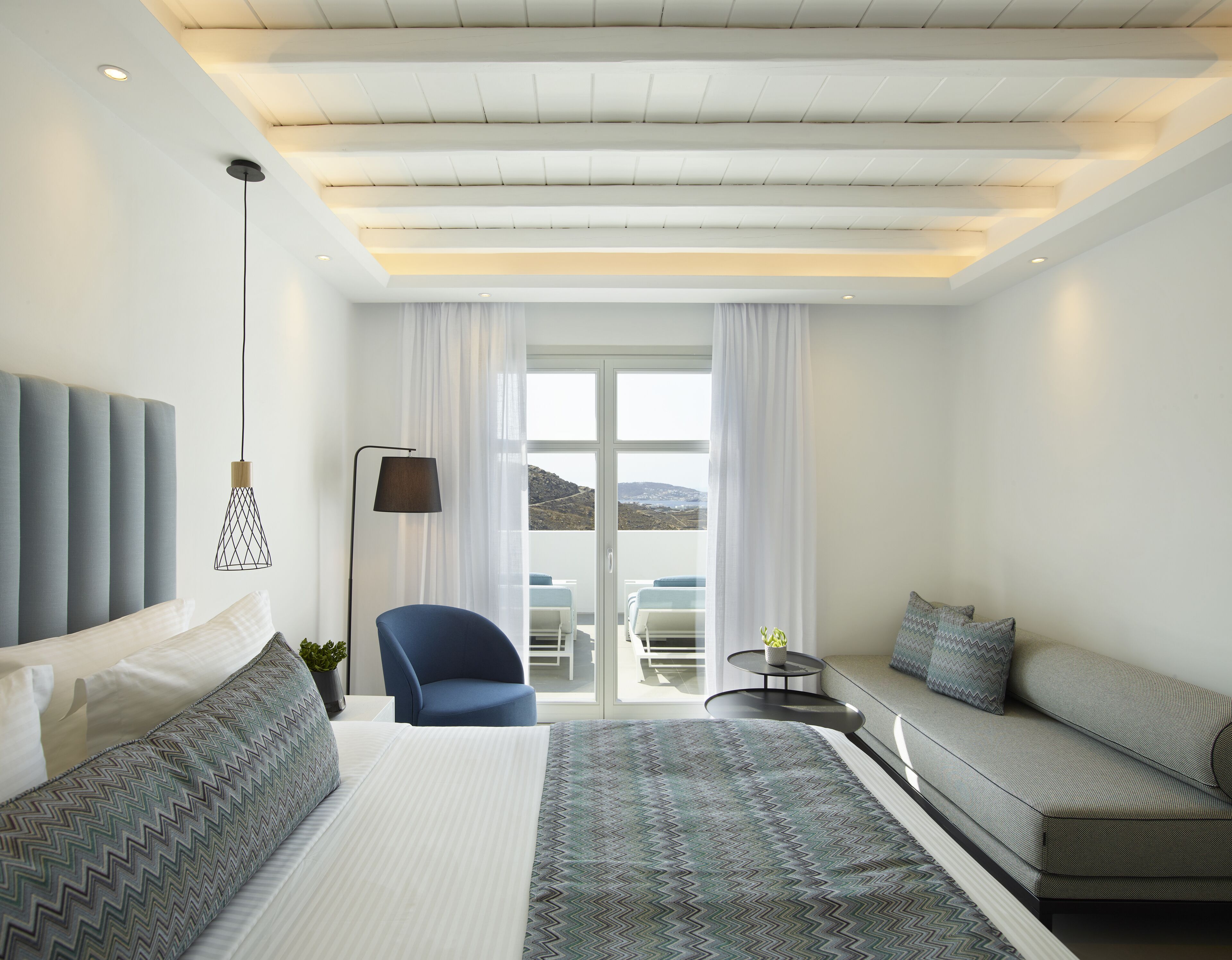 A modern hotel room with a large bed, a sofa, a chair, and a balcony with a view.