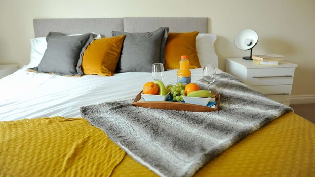 Un lit bien fait, avec une assiette de fruits, des verres et une bouteille de jus d’orange.