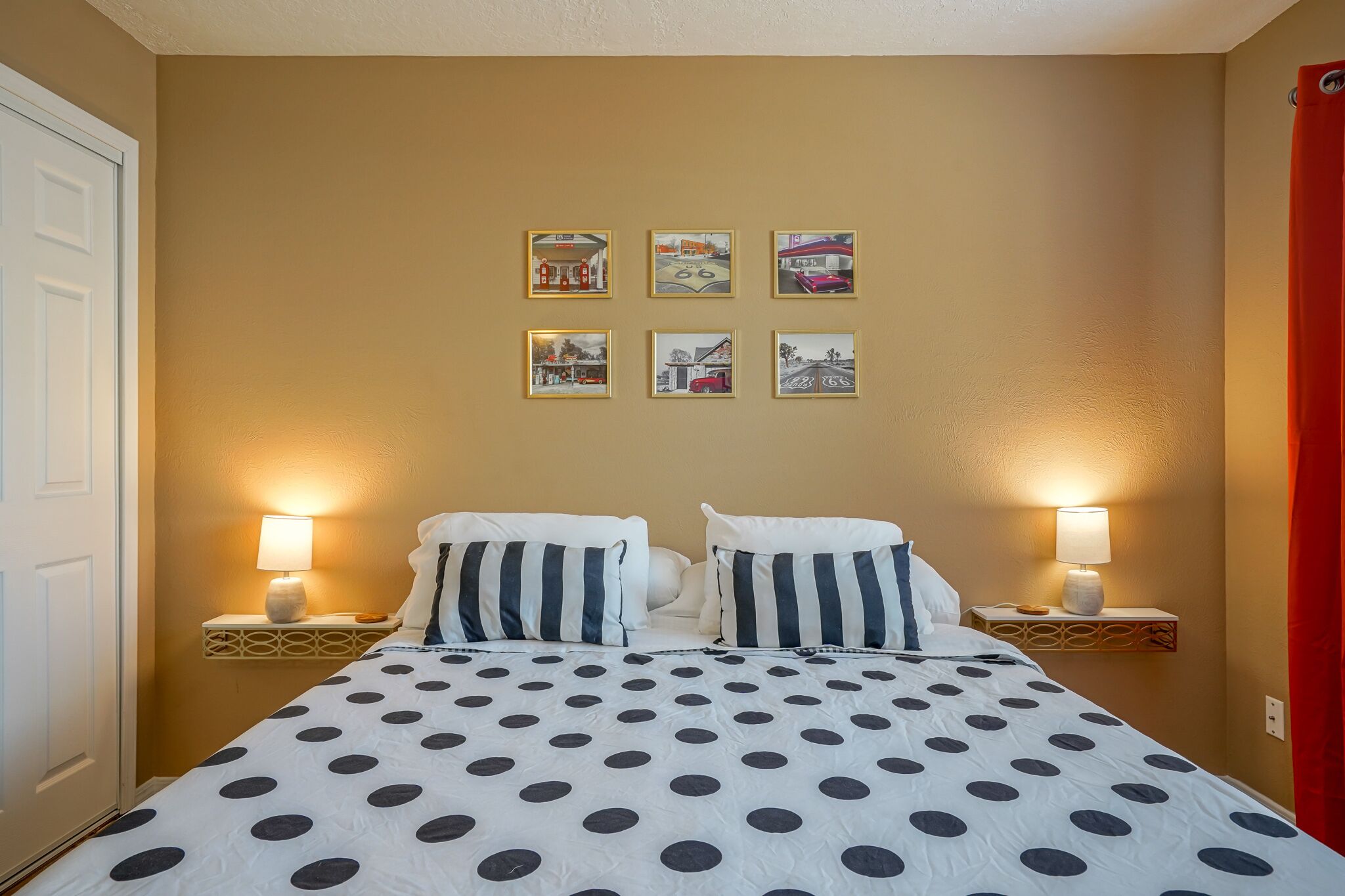 Une chambre d’hôtel avec un lit recouvert d’une courtepointe à pois, deux lampes de chevet et six tableaux encadrés accrochés au mur.