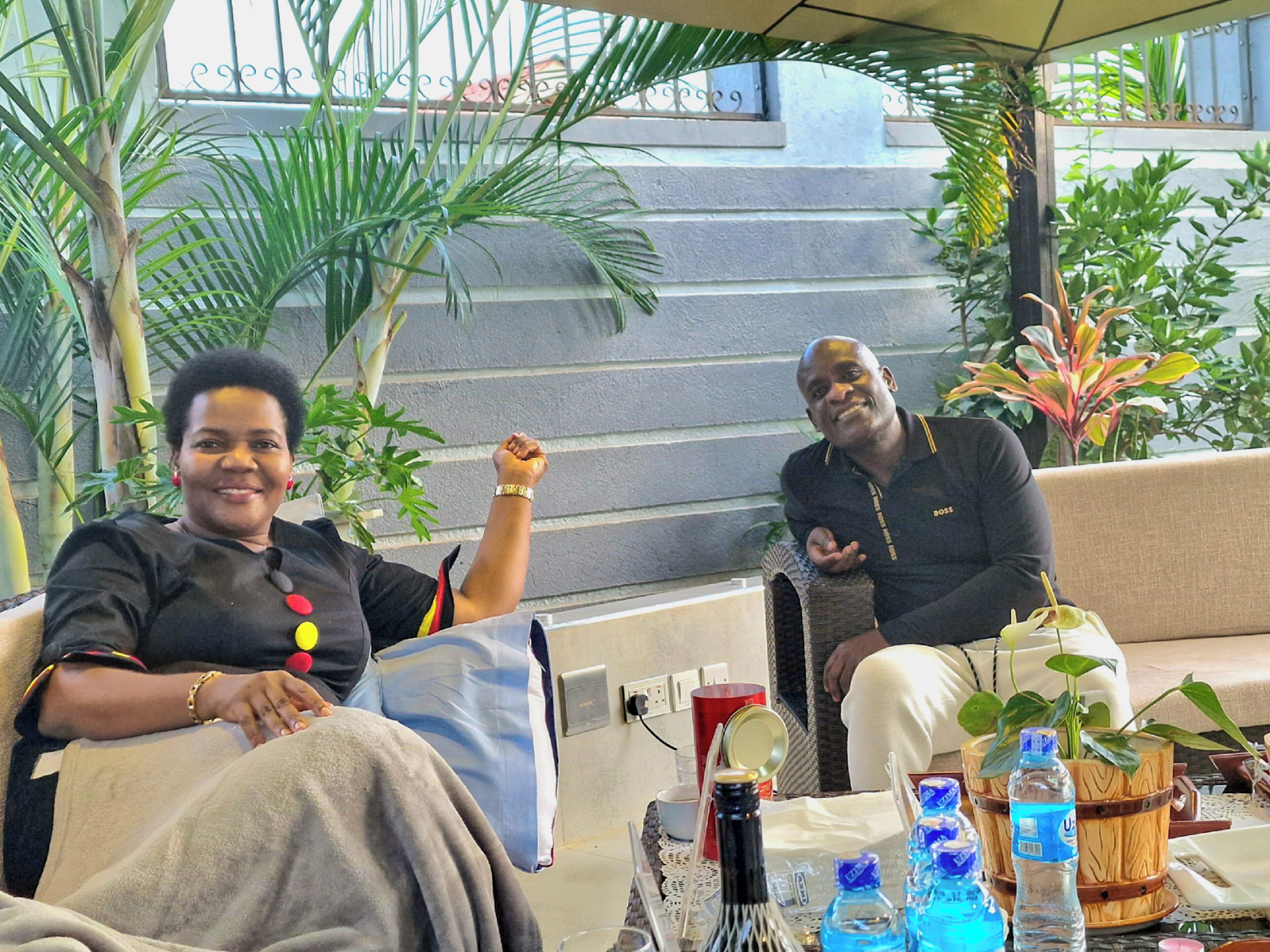Two people seated at an outdoor table with a drink and water bottles, surrounded by plants and a staircase.
