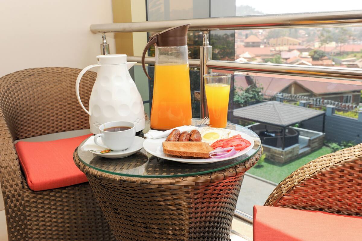 A balcony with wicker furniture, a glass table, a white pitcher, a glass of orange juice, a plate with toast, sausages, a fried egg, and a cup of coffee.