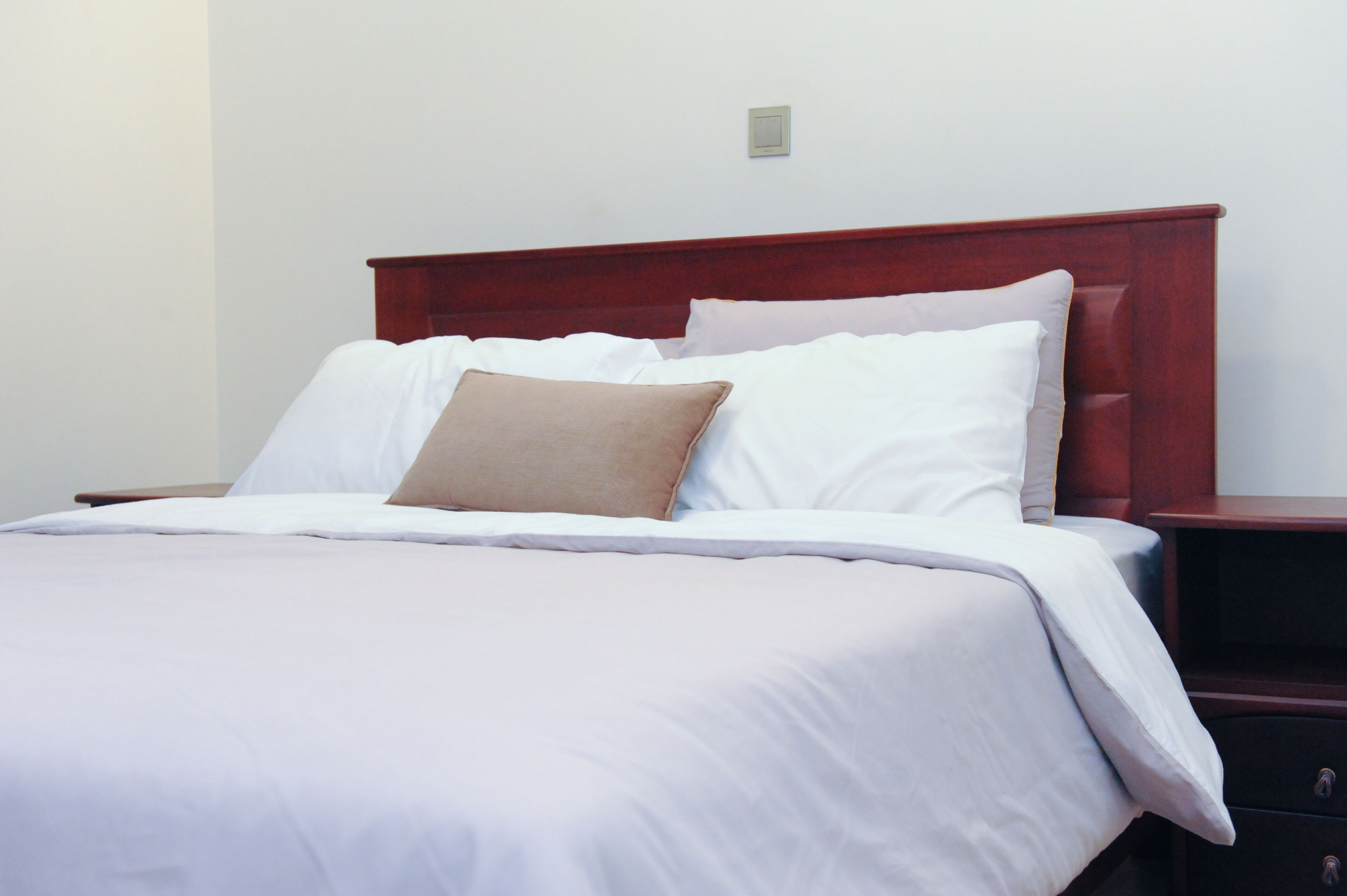 Een netjes opgemaakt bed met wit en beige beddengoed, een houten hoofdbord en een nachtkastje met lade.