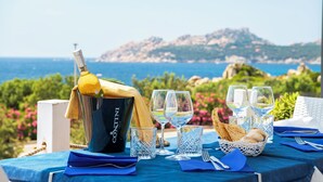 Une table dressée avec des verres à vin, une bouteille de champagne et un panier de pain, offrant une vue panoramique sur la côte.