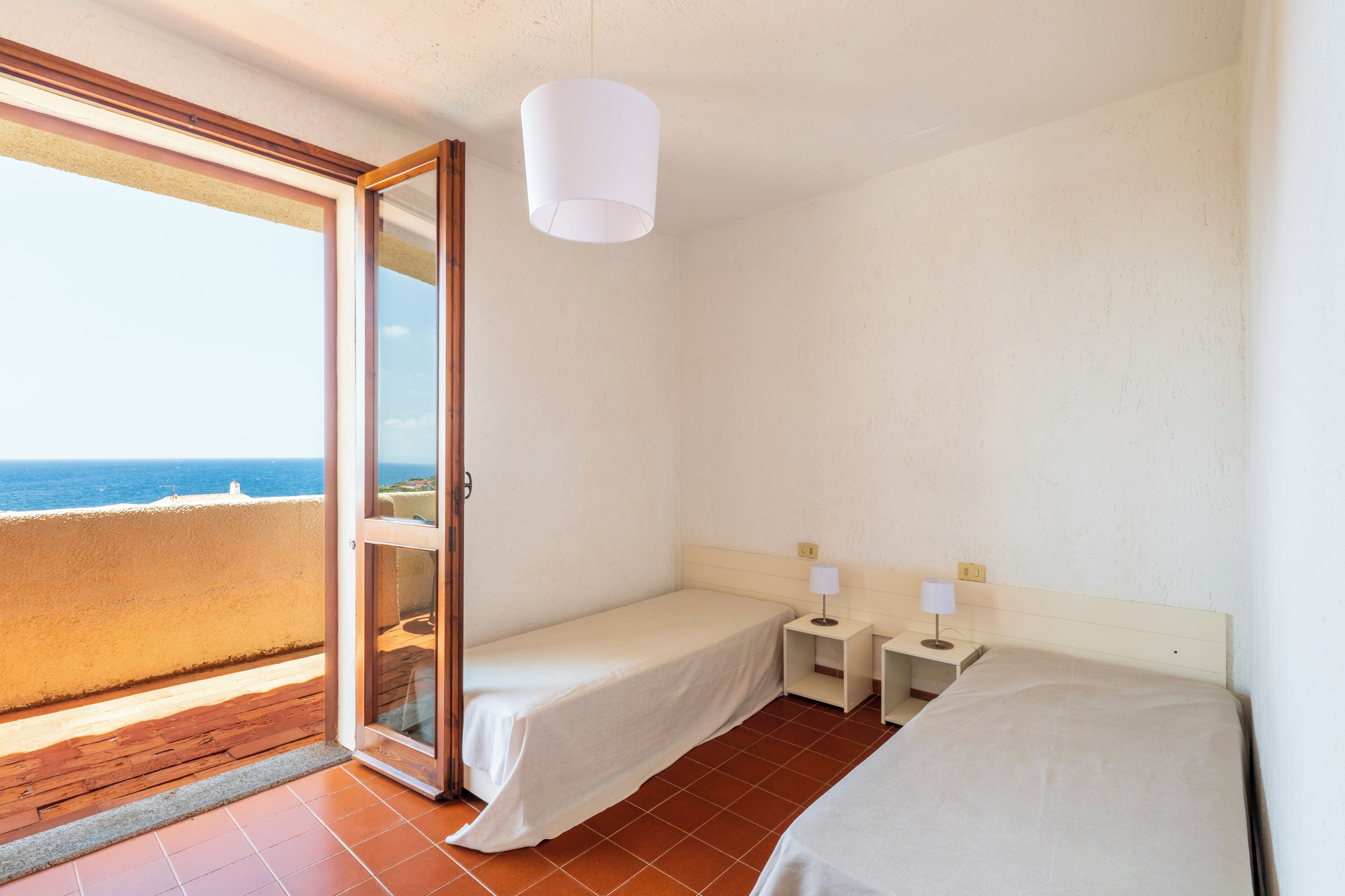 Une chambre avec deux lits, une petite table et une table de chevet. Une porte-fenêtre coulissante donne sur un balcon avec vue sur la mer.