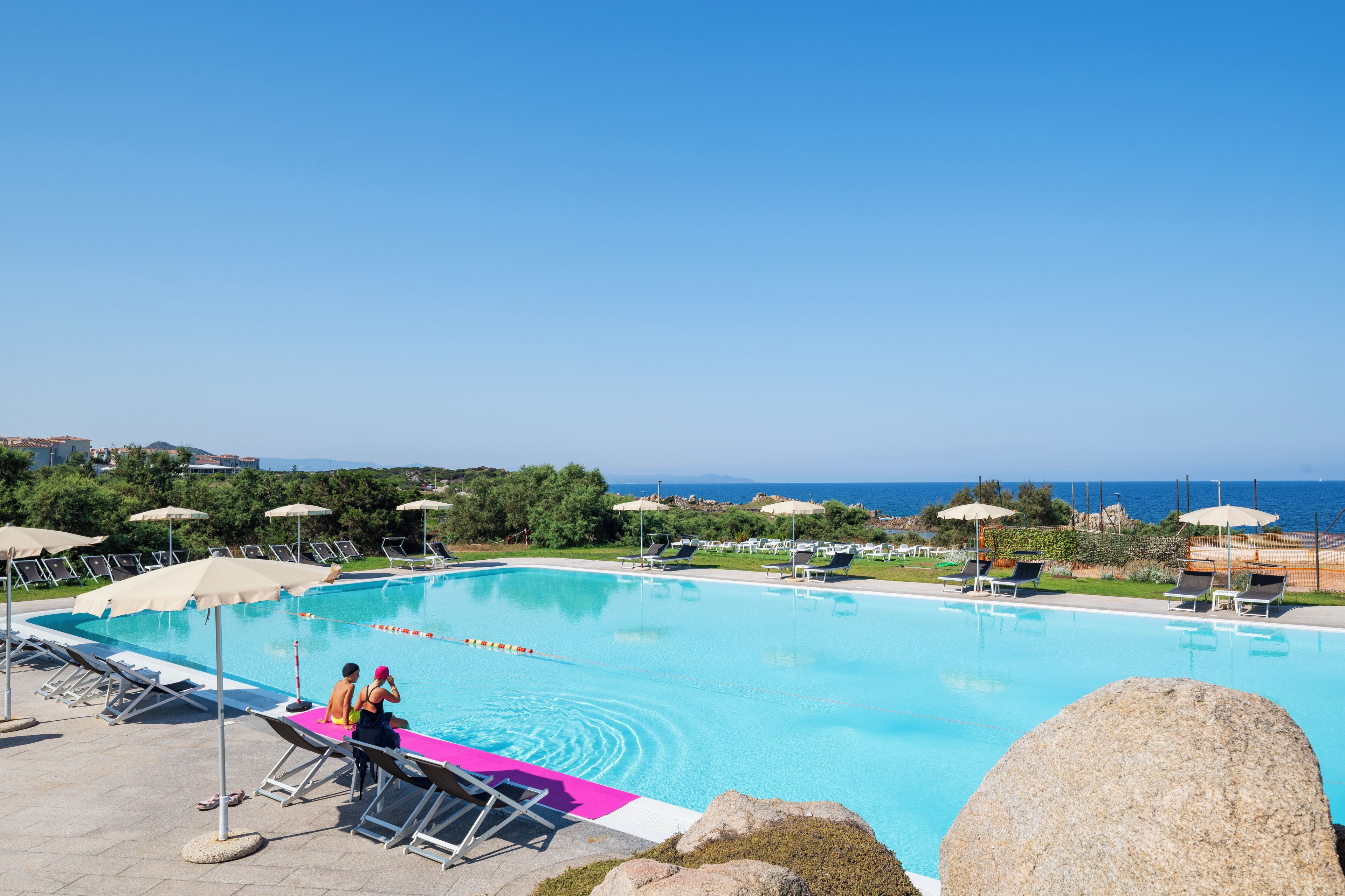 A swimming pool with lounge chairs and umbrellas, surrounded by greenery and a view of the sea.
