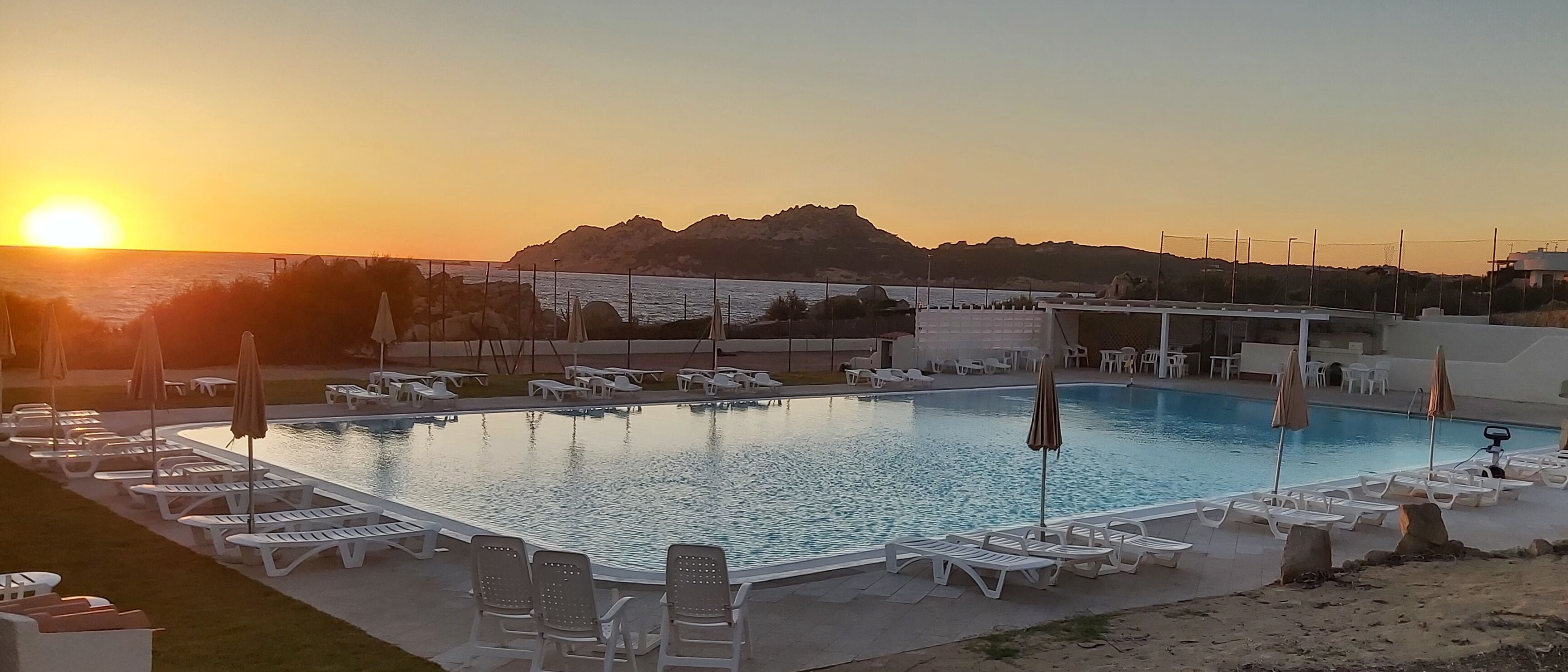 Un espace piscine agrémenté de chaises longues et de parasols, entouré d’un jardin paysager et offrant une vue sur le coucher de soleil sur la mer.