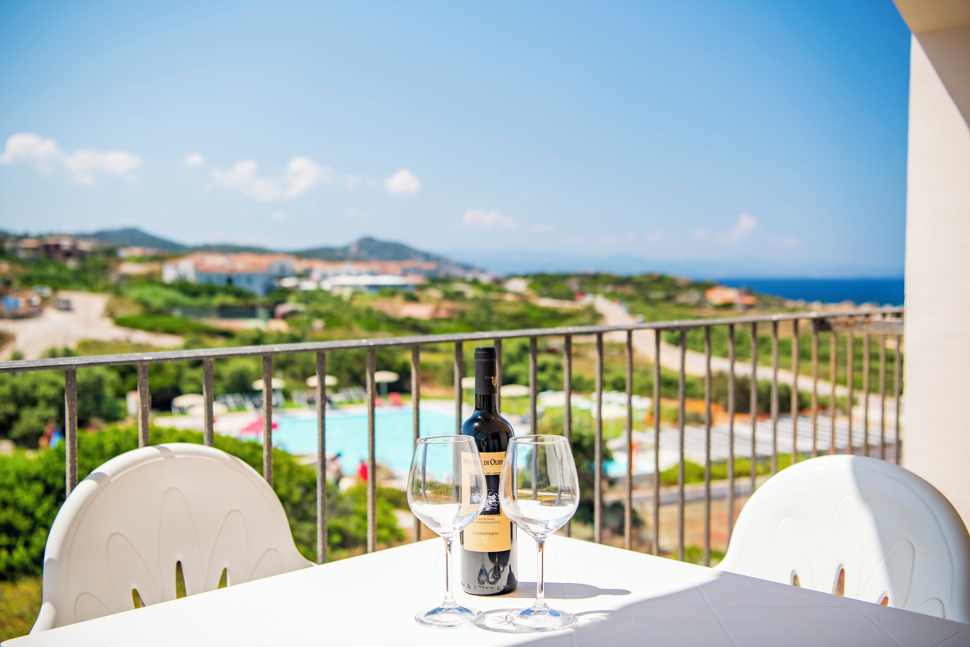 Una mesa con una botella de vino y dos copas en un balcón con vistas a un paisaje pintoresco que incluye una piscina y edificios a la distancia.