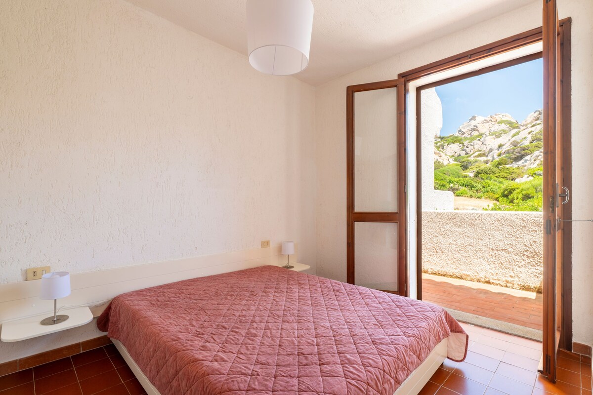 A bedroom with a bed, a nightstand, a lamp, and a sliding glass door leading to a balcony with a view of rocky terrain.