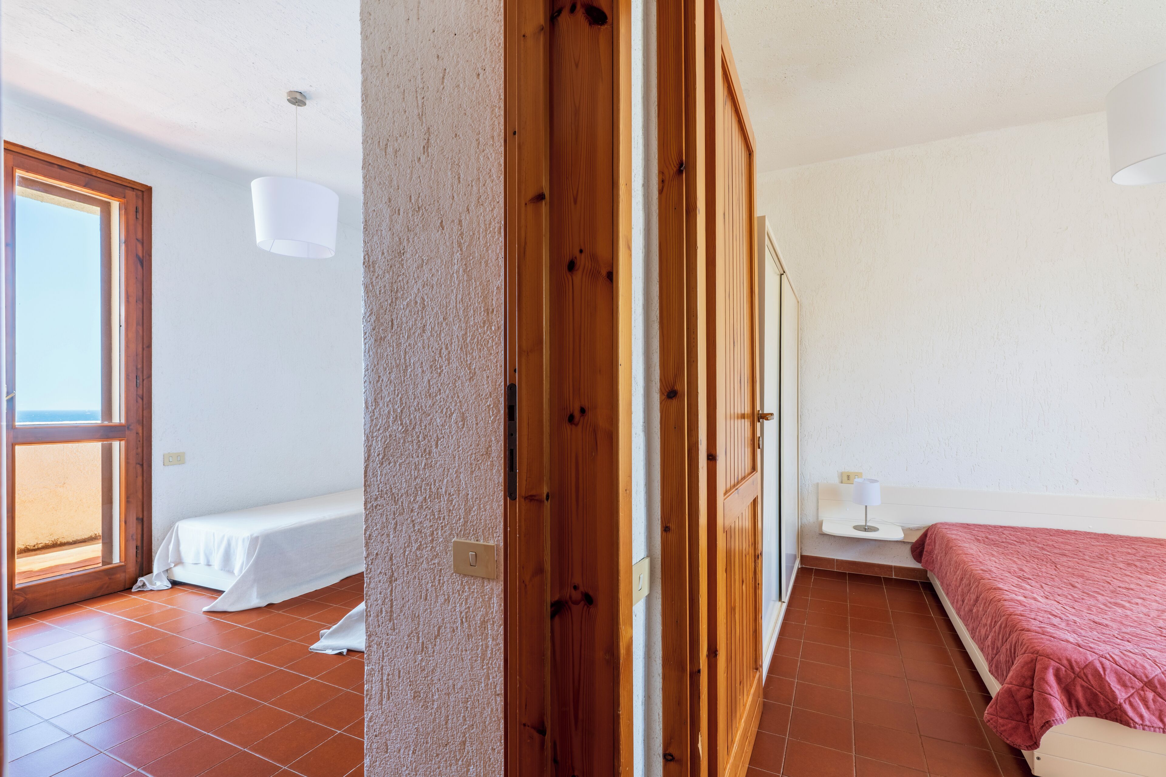 Un dormitorio con puerta de madera, ventana, cama con manta roja y mesita de noche con lámpara.