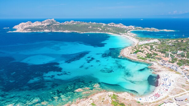An aerial view of a coastal area with a beach, clear blue waters, and a rocky island.
