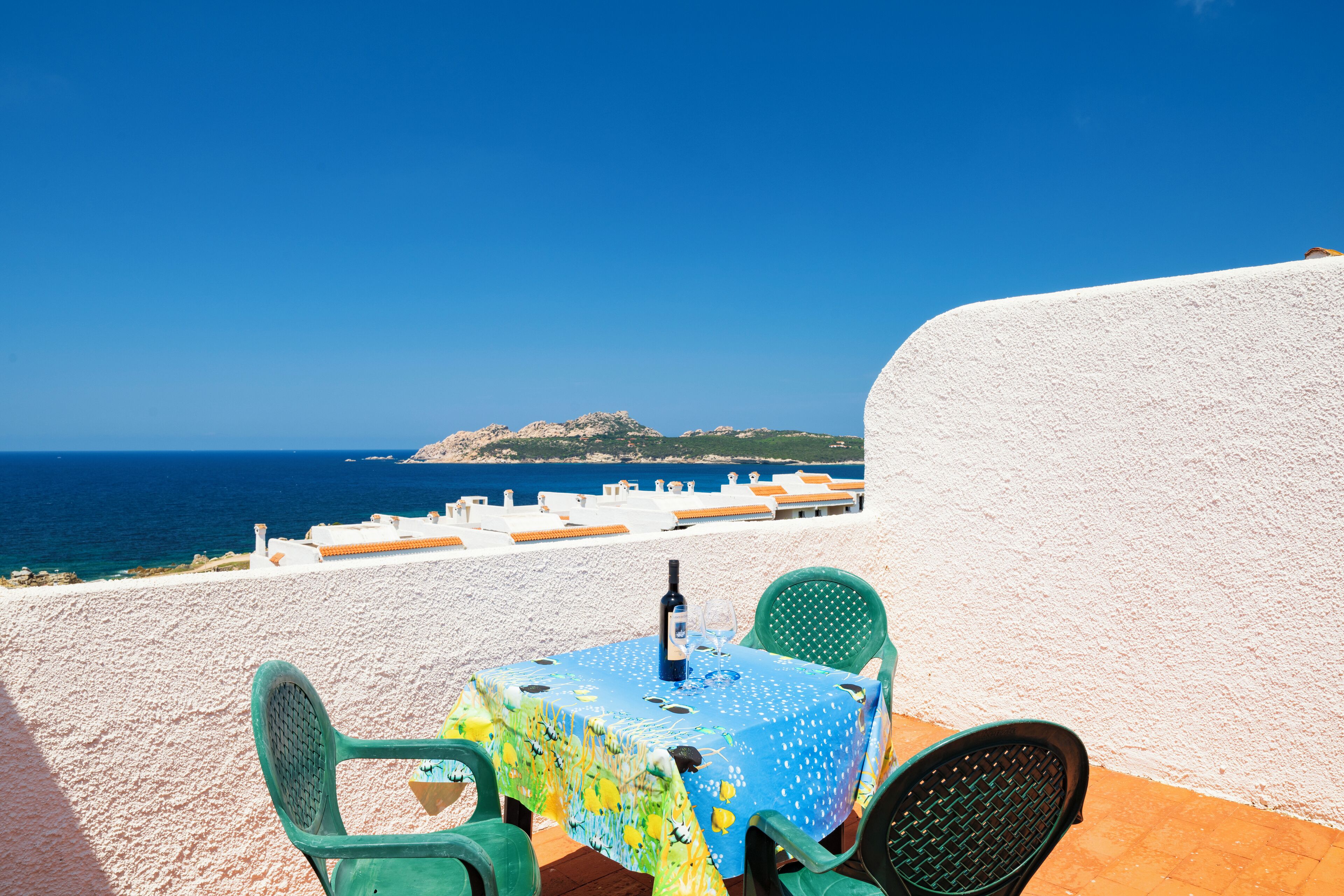 A table with a blue tablecloth and a bottle, set against a white wall with a view of the sea and a distant island.