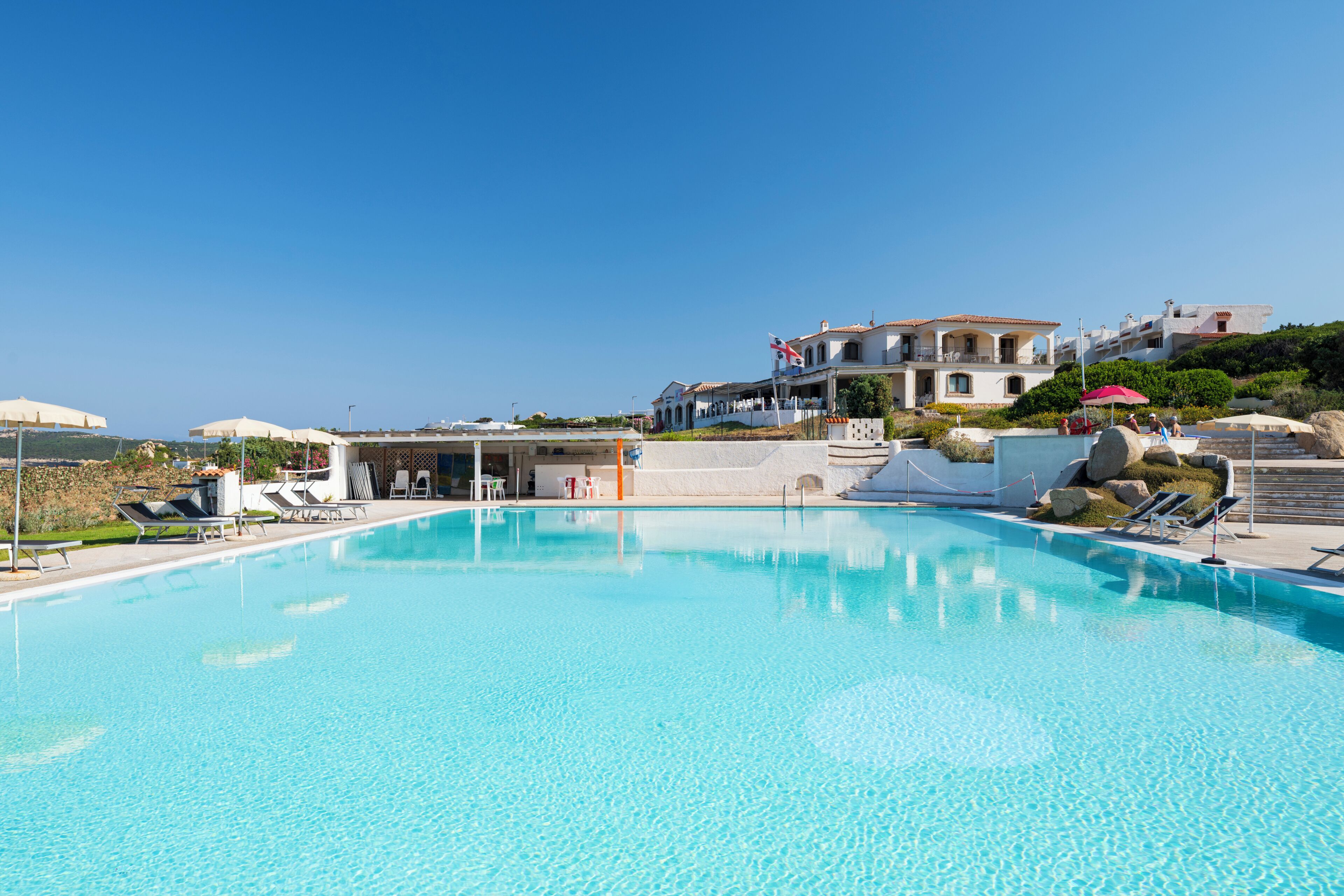 Une piscine d’un bleu éclatant, dotée de chaises longues et de parasols, entourée d’un bâtiment arborant un drapeau, et offrant une vue sur la mer.