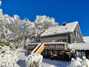 Una casa de dos pisos con un porche cubierto y una terraza de madera, rodeada de árboles cubiertos de nieve y un cielo azul despejado.