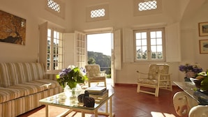 Ein geräumiges Wohnzimmer mit einem gestreiften Sofa, einem Glastisch und zwei weißen Sesseln. Große Fenster sorgen für natürliches Licht und einen Ausblick ins Freie.