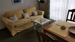 Un salon avec un canapé beige, une petite table d’appoint blanche, une table à manger en bois surmontée d’un set de table en fibres tressées et un sol à motifs.