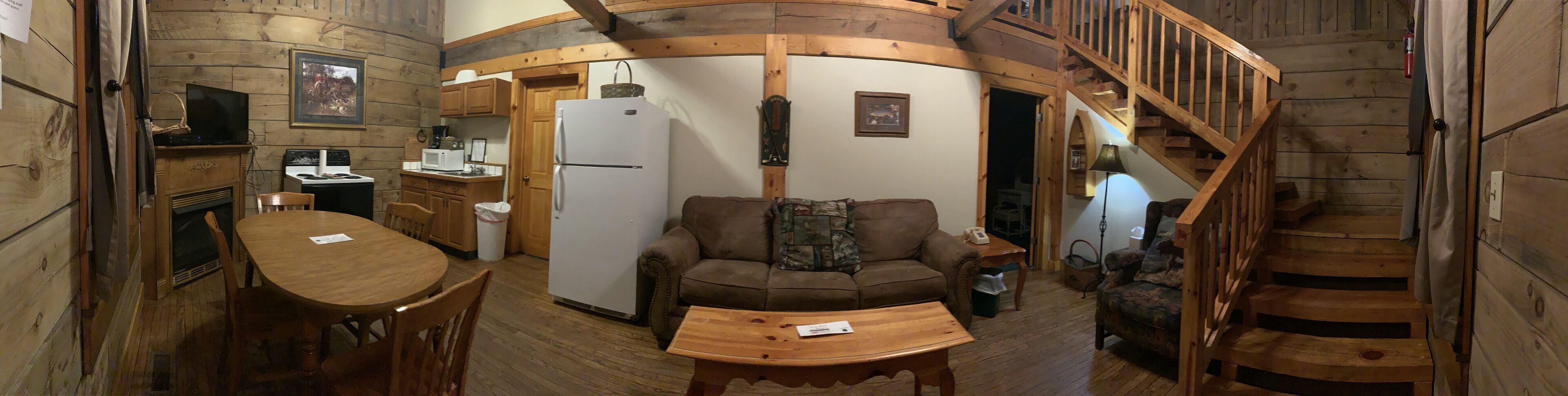 A cozy cabin interior with a wooden dining table, a sofa, a kitchen area with a refrigerator and microwave, and a spiral staircase.