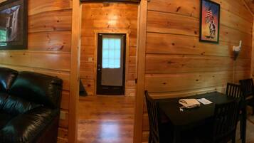 A wooden cabin interior with a leather couch, a dining table with chairs, and a door with a window.