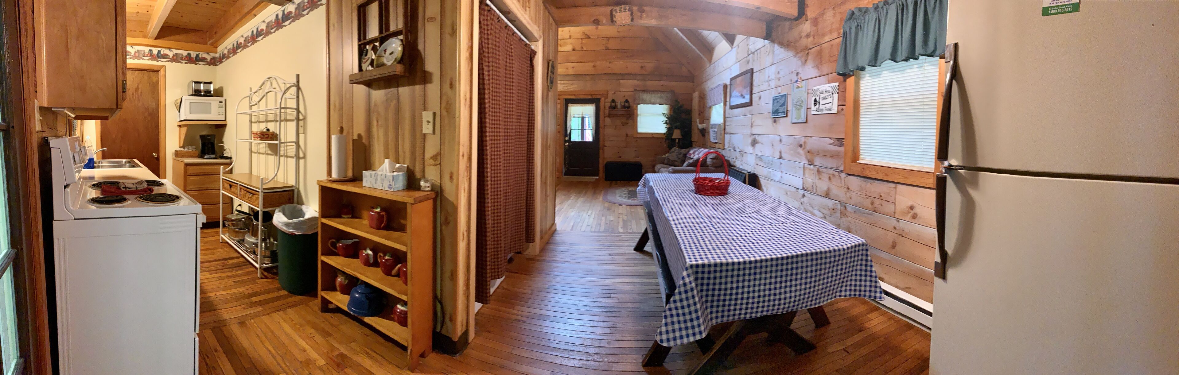 Une pièce étroite, aménagée comme une cabane en bois, avec un coin cuisine sur la gauche, une table à manger recouverte d’une nappe à carreaux au centre et un réfrigérateur sur la droite.