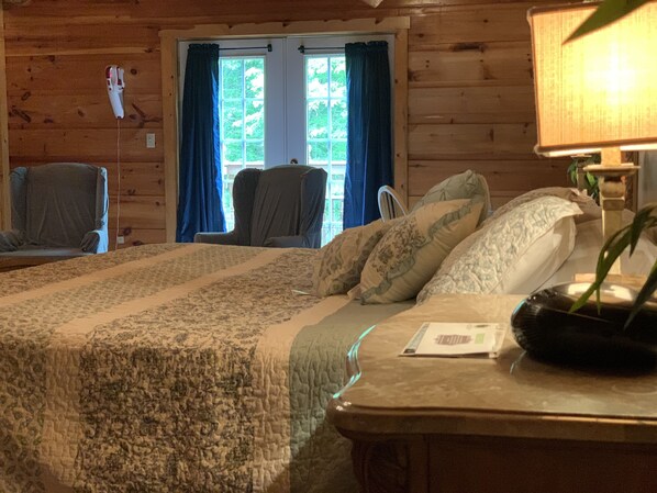 Una habitación tipo cabaña de madera con una cama, una silla, una lámpara y un ventanal con cortinas.