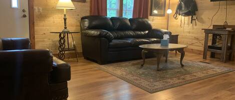 A cozy living room with a leather sofa, a coffee table, and a rug. There is a lamp, a TV mounted on the wall, and a window with curtains.