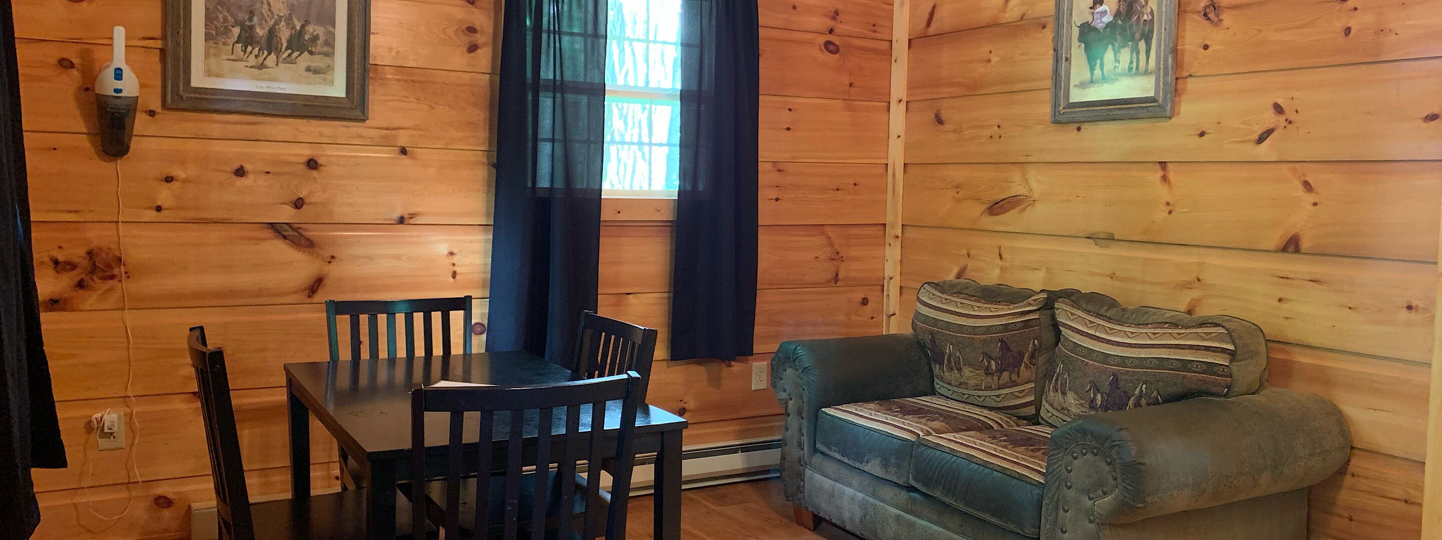Interior d'una cabana de fusta amb una taula de dinar, dues cadires, un sofà amb coixins estampats i dos quadres a les parets.