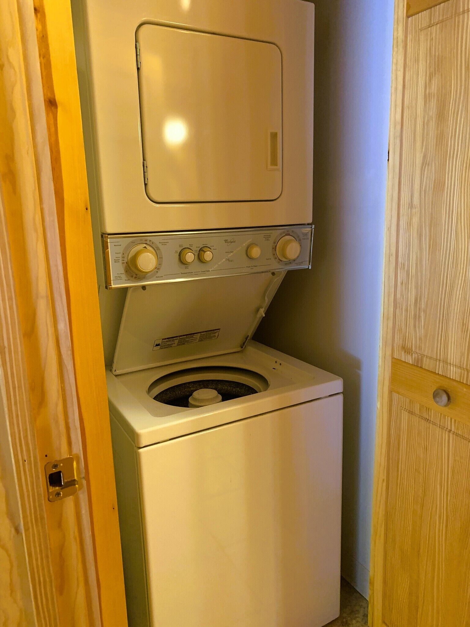 A compact, white washer and dryer unit with a front-loading design, located in a small, enclosed laundry room with wooden doors.