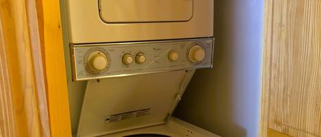 A compact, white washer and dryer unit with a front-loading design, located in a small, enclosed laundry room with wooden doors.