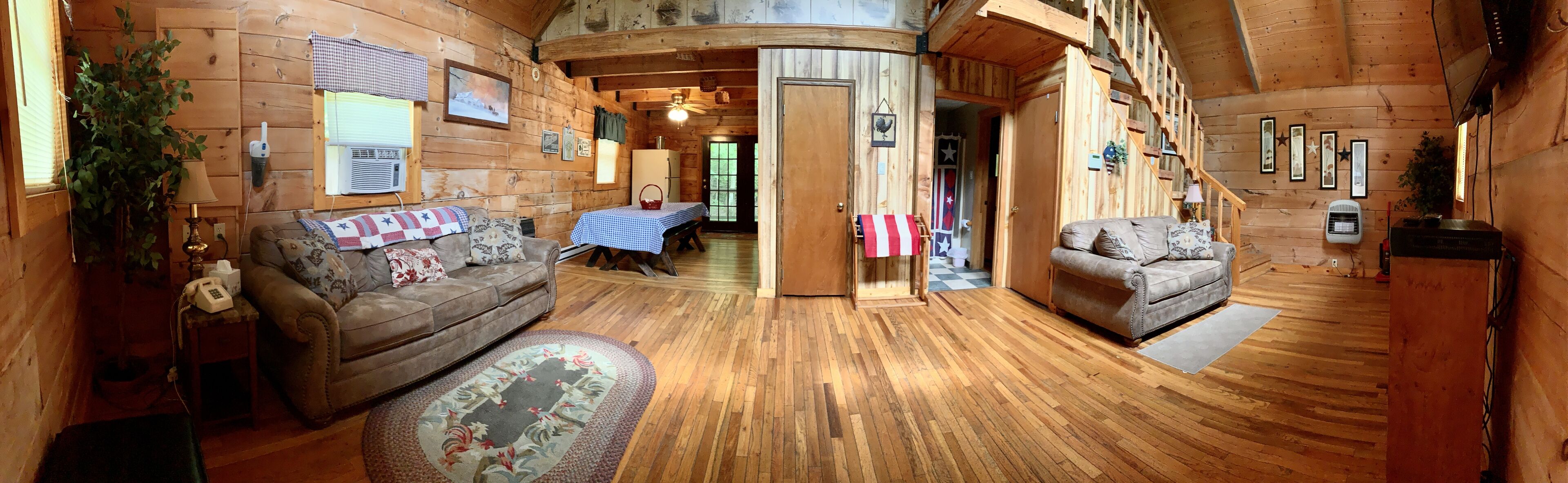A cozy cabin interior with wooden walls, a wooden floor, a sofa, a dining table with a checkered tablecloth, and a flag.