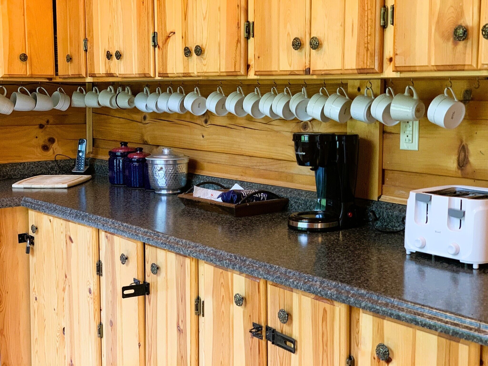 Eine Küche mit Holzausstattung, einer Kaffeemaschine, einem Toaster und einer Reihe von Tassen, die an der Wand hängen.