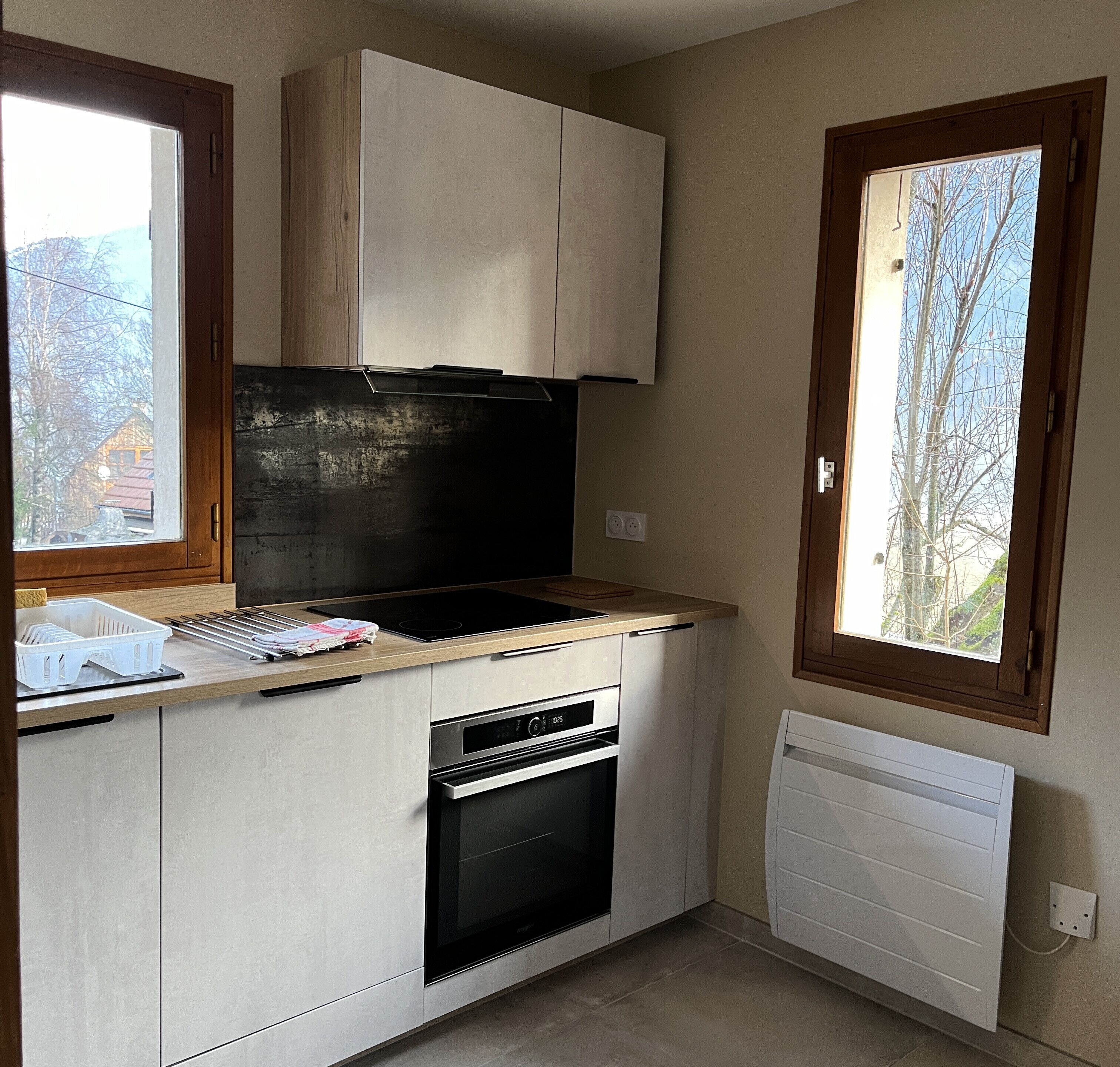 Une cuisine compacte équipée d’un four encastré, d’une plaque de cuisson et d’une fenêtre donnant sur l’extérieur.