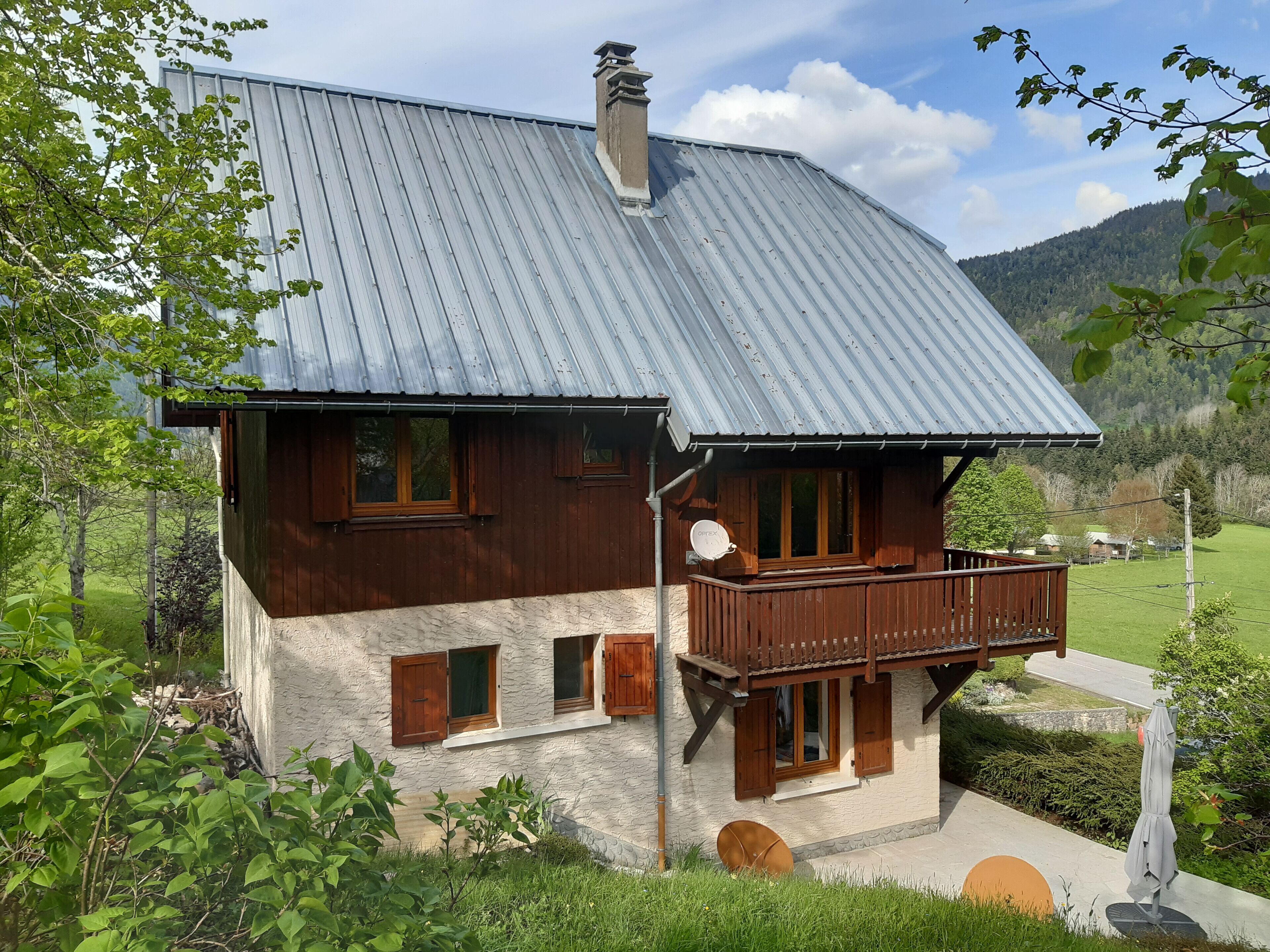 A two-story wooden house with a balcony, surrounded by greenery and a mountainous landscape.