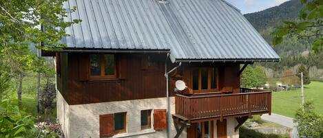 A two-story wooden house with a balcony, surrounded by greenery and a mountainous landscape.