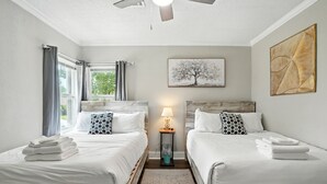 Un dormitorio con dos camas, un ventilador de techo, una ventana con cortinas, una lámpara y un cuadro en la pared.