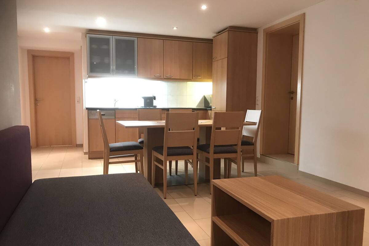 Un espacio habitable compacto con kitchenette, una zona de comedor con cuatro sillas, un sofá y una pequeña mesa de madera.