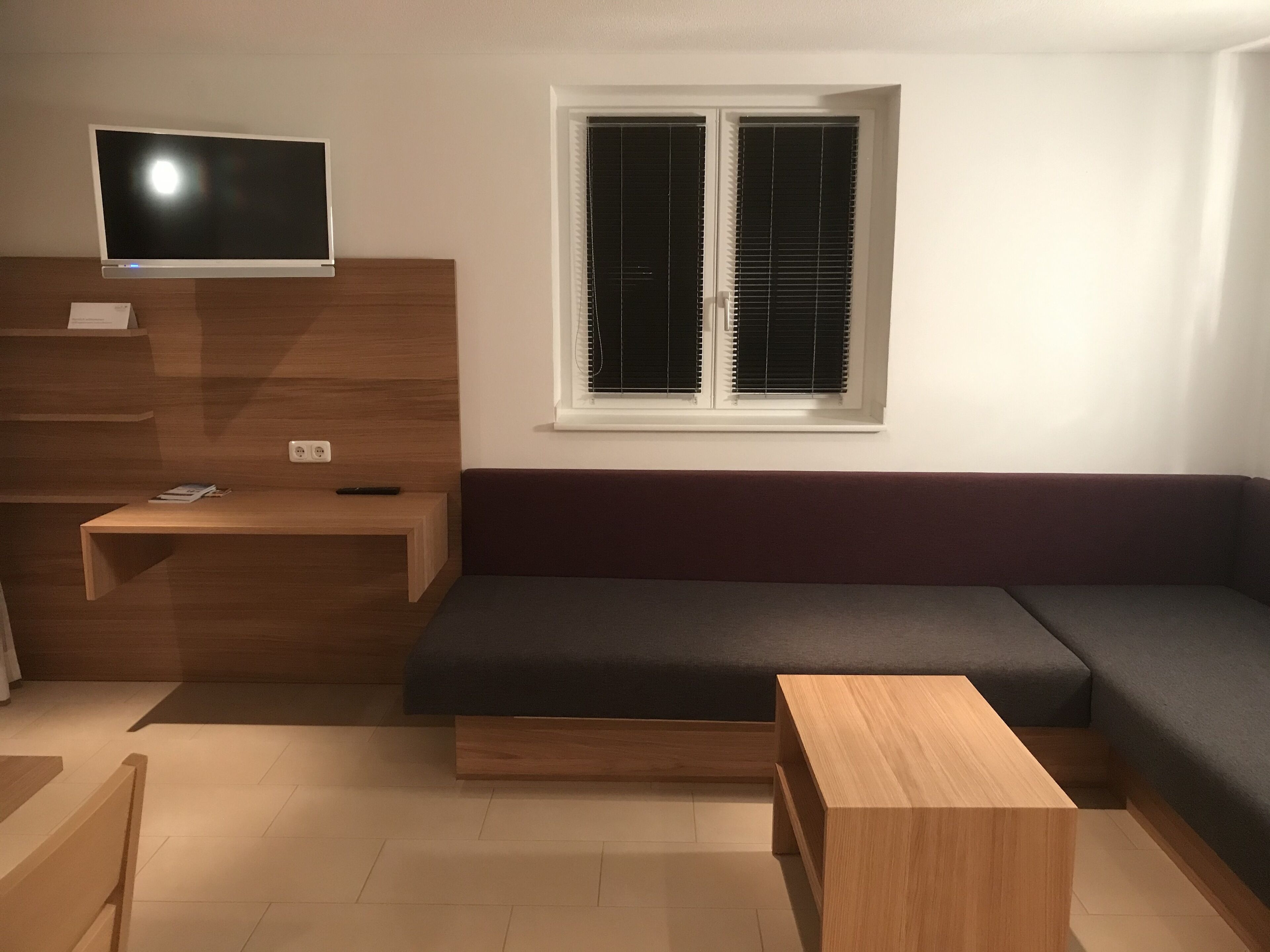 A modern living room with a dark-colored sofa, a wooden TV stand, a small wooden table, and a window with blinds.