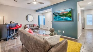 A modern living room with a grey sofa, red bar stools, a round mirror, a flat-screen TV, and a yellow rug. - Sleek Modern 4BR Near Fort Bliss (El Paso)