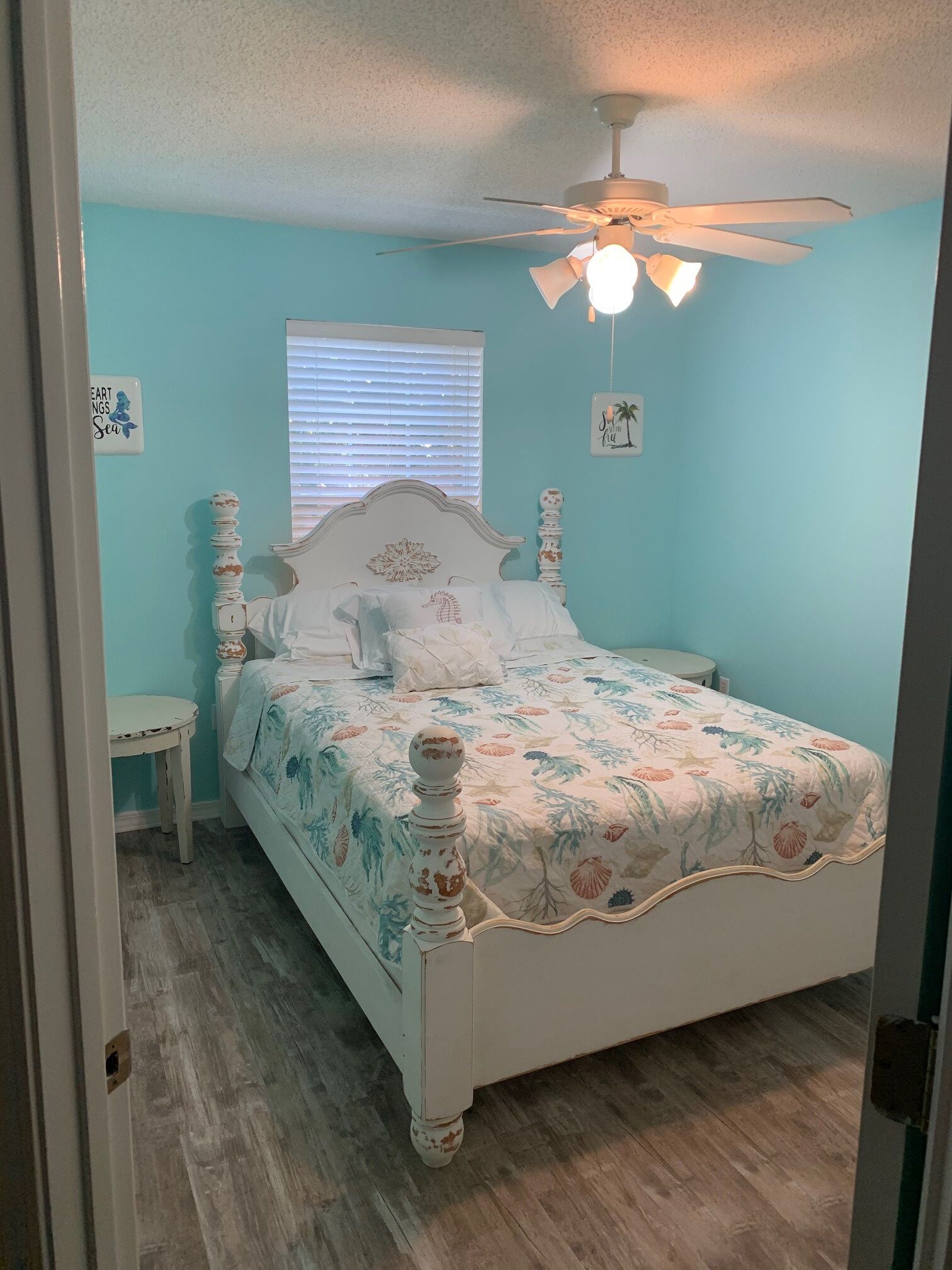 Une chambre avec un lit, un ventilateur de plafond, une fenêtre avec des stores et une petite table.