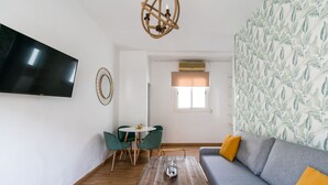 Een woonkamer met een flatscreen televisie, een ronde tafel met stoelen, een bank met kussens, een houten salontafel, een kroonluchter en een raam met zonwering.