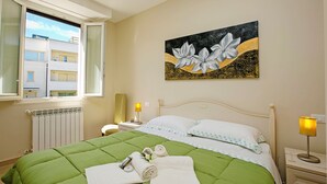 A hotel room with a green bedspread, a painting of flowers on the wall, a window with a view of buildings, and a bedside table with a lamp. - Villa Mimosa - Apt 2 (Desenzano del Garda)