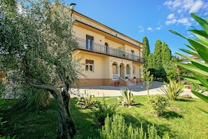 A two-story building with a balcony, surrounded by greenery and trees.