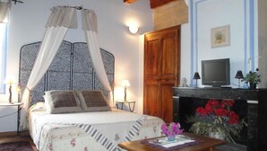 A bedroom with a four-poster bed, a television, a fireplace with red flowers, and a small table with a vase.