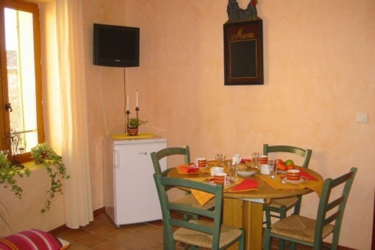 A dining area with a table set for four, a small refrigerator, a television mounted on the wall, and a decorative item above the table.