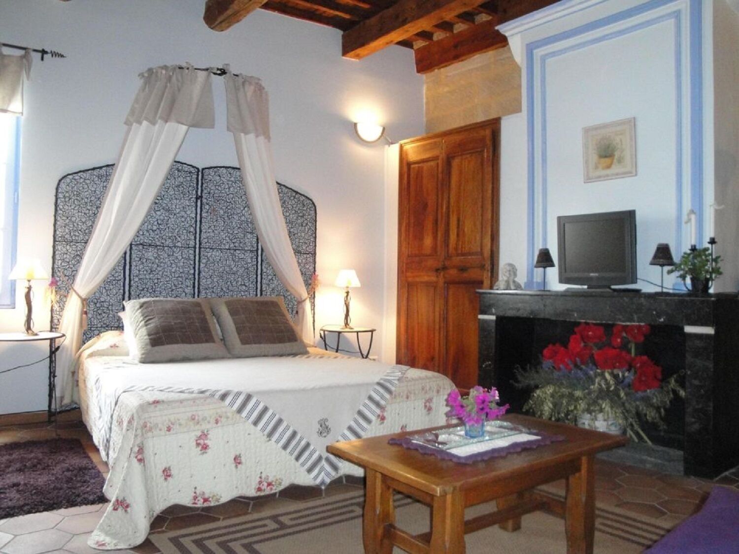 A bedroom with a four-poster bed, a television, a fireplace with red flowers, and a small table with a vase.